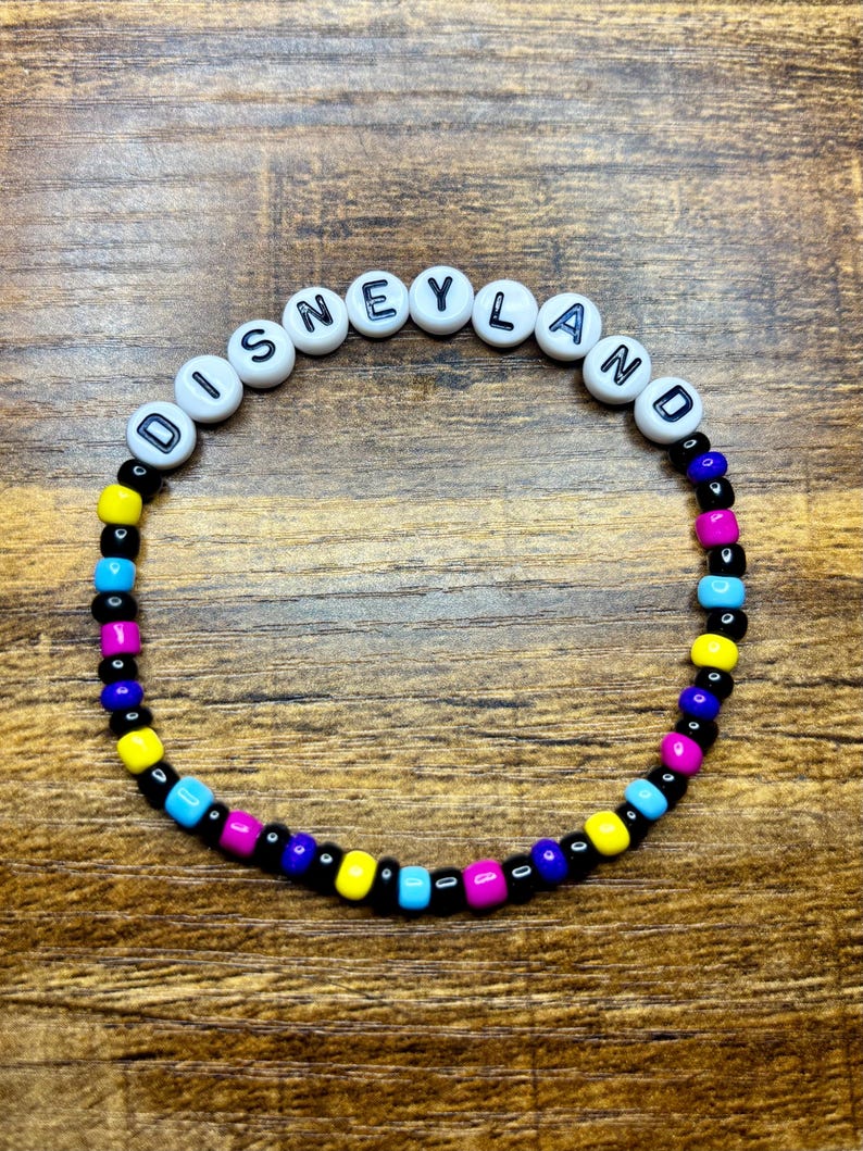 May include: A beaded bracelet with the word "DISNEYLAND" spelled out in white letter beads. The bracelet features a colorful pattern of black, yellow, blue, and pink beads, creating a vibrant accessory.