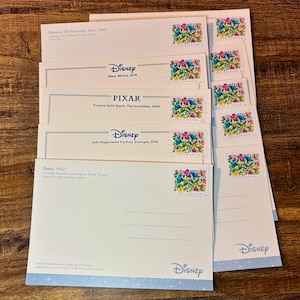 May include: A collection of Disney-themed postcards and stamps. The postcards feature titles like "Fantasia," "Moana," "The Incredibles," and "Zootopia." Each postcard has a Disney logo and is paired with a colorful floral stamp. The background is a wooden surface.