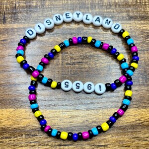 May include: Two beaded bracelets. The top bracelet spells "DISNEYLAND" in white letter beads. The bottom bracelet spells "1955" in white letter beads. Both bracelets have colorful beads in blue, yellow, pink, and black.