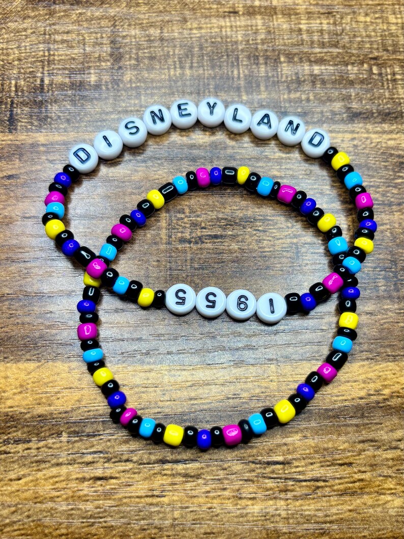 May include: Two beaded bracelets. The top bracelet spells "DISNEYLAND" in white letter beads. The bottom bracelet spells "1955" in white letter beads. Both bracelets have colorful beads in blue, yellow, pink, and black.