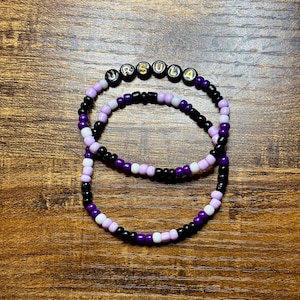 May include: Two beaded bracelets with the name "URSULA" spelled out in gold letters on black beads. The bracelets feature a color scheme of black, purple, and white beads, arranged in a pattern. The bracelets are displayed on a wooden surface.