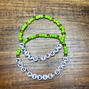 May include: Two beaded bracelets. The top bracelet is lime green, and the bottom bracelet is lime green with white letter beads that spell out "FAITH TRUST AND PIXIE DUST". The bracelets are on a wooden surface.