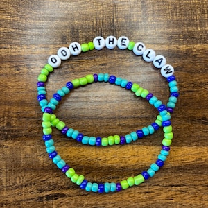 May include: Two beaded bracelets. One bracelet spells out "OOH THE CLAW" with white letter beads. The other bracelets are made of blue, green, and purple beads. The bracelets are on a brown wooden surface.