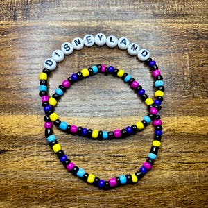 May include: Two beaded bracelets. The top bracelet has white beads spelling out "DISNEYLAND". The bottom bracelet and the letters on the top bracelet are black, yellow, blue, and pink. The bracelets are on a wooden surface.