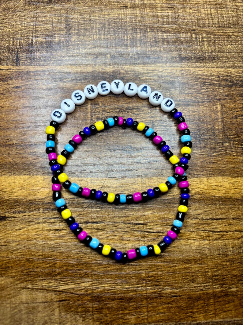 May include: Two beaded bracelets. The top bracelet has white beads spelling out "DISNEYLAND". The bottom bracelet and the letters on the top bracelet are black, yellow, blue, and pink. The bracelets are on a wooden surface.