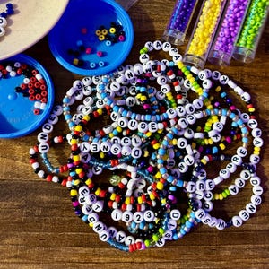 May include: A close-up of a pile of colorful beaded bracelets with black letter beads spelling out words like "MOUSE", "WISH", "SNEY", "TRE", "OZMA", "KAPO", "UNDER", and "923".