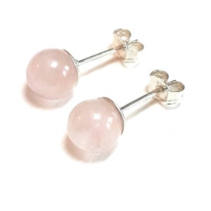 Può includere: Un paio di orecchini a lobo con pietre sferiche di quarzo rosa pallido. Le pietre sono montate su perni d'argento con chiusure a farfalla, su sfondo bianco. Gli orecchini sono semplici ed eleganti.