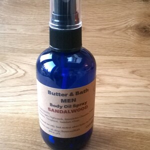 Op de afbeelding: Een blauwe glazen fles met een zwarte spuitdop en een bruin etiket met de tekst "Butter & Bath MEN Body Oil Spray SANDALWOOD".
