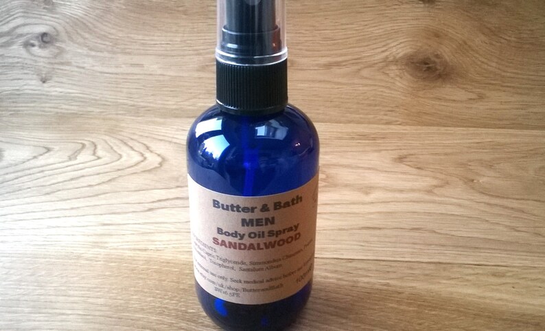 Op de afbeelding: Een blauwe glazen fles met een zwarte spuitdop en een bruin etiket met de tekst "Butter & Bath MEN Body Oil Spray SANDALWOOD".