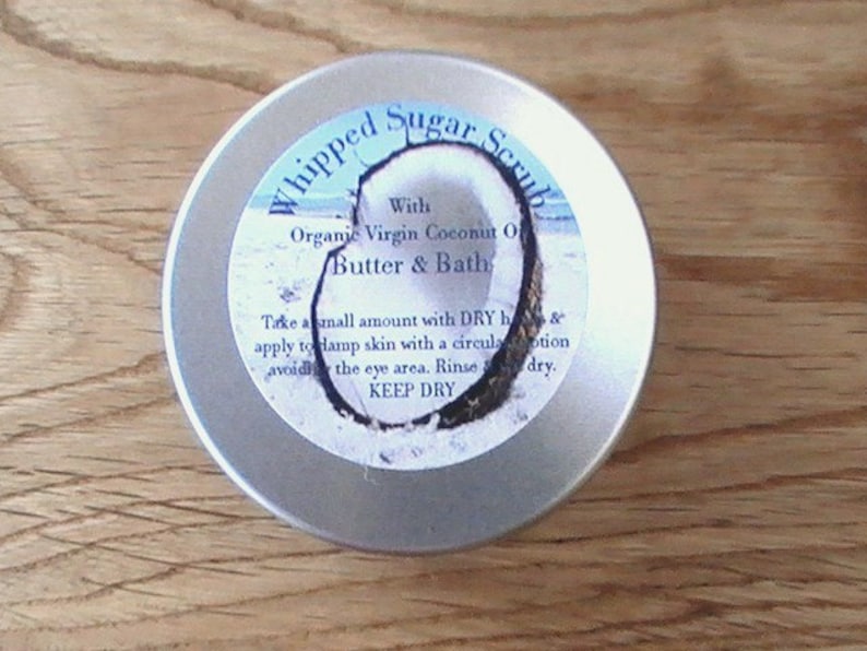 May include: A silver tin container with a white label that reads "Whipped Sugar Scrub" with the text "With Organic Virgin Coconut Oil Butter & Bath". The label also includes instructions for use: "Take a small amount with DRY body & apply to damp skin with a circular motion, avoiding the eye area. Rinse & dry. KEEP DRY."