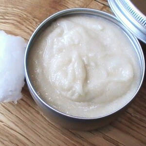 May include: A close-up of a small, silver tin filled with a white, creamy substance. The tin is sitting on a wooden surface.