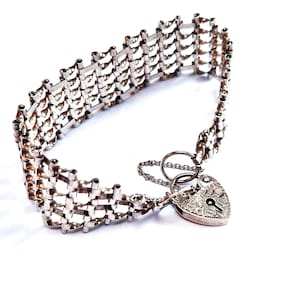 Könnte beinhalten: Ein silbernes Armband mit einem herzförmigen Vorhängeschloss. Das Armband besteht aus miteinander verbundenen rechteckigen Gliedern. Das herzförmige Vorhängeschloss hat ein eingraviertes Design und ein Schlüsselloch. Eine kleine Kette verbindet das Schloss mit dem Armband.