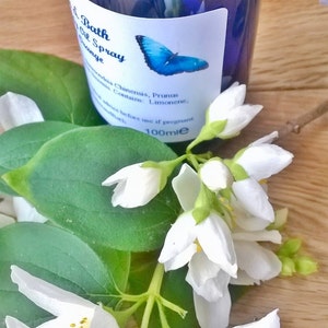 May include: A blue glass bottle with a white label that says "Bath Oil Spray Orange" and lists ingredients. The bottle is sitting on a wooden surface with white flowers.