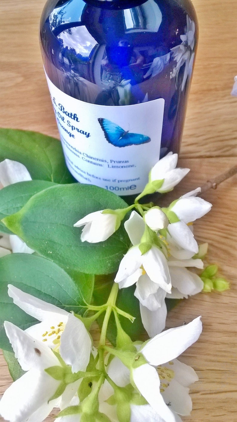 May include: A blue glass bottle with a white label that says "Bath Oil Spray Orange" and lists ingredients. The bottle is sitting on a wooden surface with white flowers.
