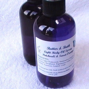 May include: Two purple glass bottles with white labels. The label on the front bottle reads "Butter & Bath Light Body Oil Spray Patchouli & Sweet Orange". The label also includes a list of ingredients.