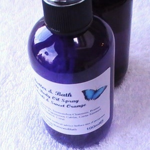 May include: A purple glass bottle with a black spray top and a white label. The label says "Butter & Bath Body Oil Spray Sweet Orange" and has a blue butterfly illustration. The bottle contains 100 ml of liquid.