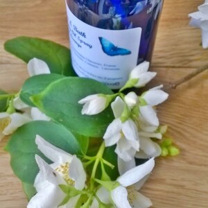 May include: A blue glass bottle with a white label that says "Bath & Body Spray Orange" and a blue butterfly graphic. The bottle is sitting on a wooden surface with white jasmine flowers.