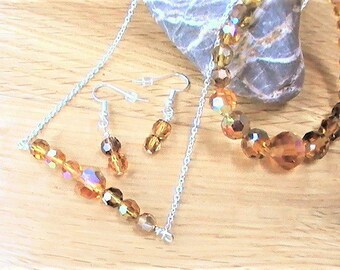 Op de afbeelding: Een set amber kristallen sieraden met een ketting en oorbellen. De ketting is een eenvoudige ketting met een rij facetgeslepen amber kristallen. De oorbellen zijn hangende oorbellen met een enkel facetgeslepen amber kristal.