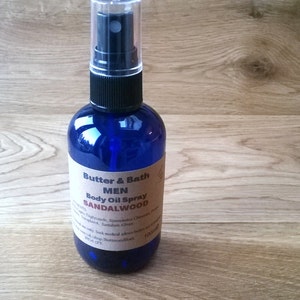 Op de afbeelding: Een blauwe glazen fles met een zwarte spuitdop. De fles is gelabeld met "Butter & Bath Men Body Oil Spray Sandalwood".