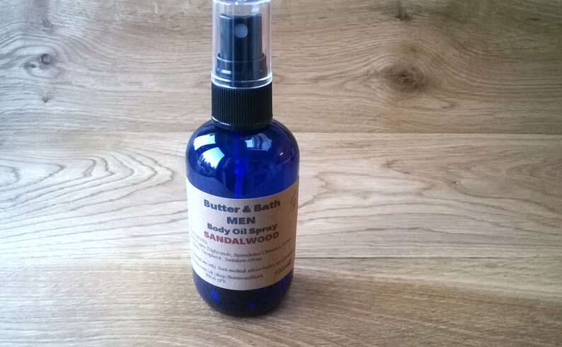 Op de afbeelding: Een blauwe glazen fles met een zwarte spuitdop. De fles is gelabeld met "Butter & Bath Men Body Oil Spray Sandalwood".