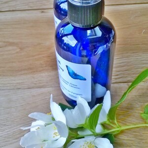 May include: Two blue glass spray bottles with black caps and white labels. The labels have a butterfly design and list the ingredients:  "Orange, Lemon,  Lime,  Bergamot,  Clary Sage,  Frankincense,  Prunus,  Limonene". The text "100ml" is also on the label.  White flowers with green leaves are in the foreground.