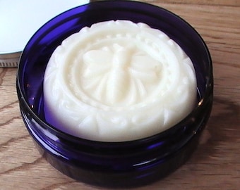 May include: A round, white, solid, unscented deodorant in a blue glass jar. The deodorant has a decorative design on the top.