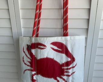 May include: White tote bag with a large red crab applique. The bag has red and white rope handles and is hanging from a wooden cleat. The crab is stitched onto the bag with white thread.