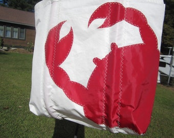 May include: White tote bag featuring a large red crab design. The crab is stitched onto the bag with white thread. The bag is made of a lightweight, possibly sailcloth material, and is suitable for carrying items.