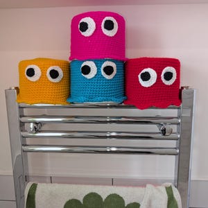 May include: Four crocheted toilet paper roll covers stacked on a chrome towel rack. The covers are in pink, blue, yellow, and red with white eyes.