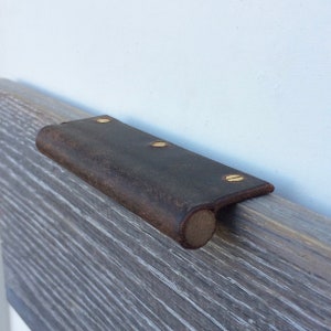 May include: A dark brown leather cabinet pull with brass screws. The pull is curved on one end and has a rectangular shape.