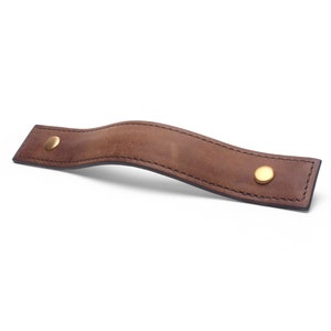 May include: A brown leather pull handle with two gold metal rivets. The handle is curved and has a stitched edge.