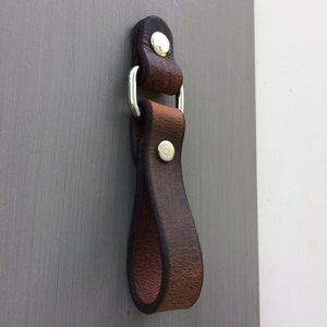 May include: A brown leather loop handle with a silver metal ring and two silver metal rivets. The handle is designed to be attached to a bag or other object.