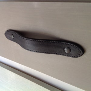May include: Black leather drawer pull with two silver rivets. The pull is curved and has a stitched edge.