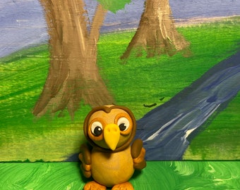 May include: A brown and yellow owl figurine with large eyes, a yellow beak, and orange feet. The owl is standing on a green painted surface with a blue stream painted in the background.