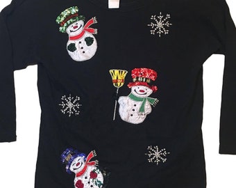 Op de afbeelding: Zwart longsleeve shirt met drie sneeuwpop-applicaties. De sneeuwpoppen zijn versierd met pailletten en kralen. Het shirt heeft ook sneeuwvlok-applicaties.