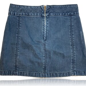 Pu&ograve; includere: Una gonna di jeans blu con una chiusura a zip sul retro. La gonna ha un taglio dritto e un orlo leggermente svasato.