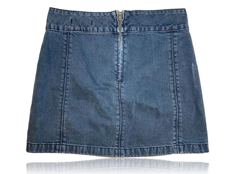 Pu&ograve; includere: Una gonna di jeans blu con una chiusura a zip sul retro. La gonna ha un taglio dritto e un orlo leggermente svasato.