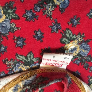 May include: A white tag with black text that reads "BONGO", "EUGENE MONTESANO", "SIZE M", and "STYLE 1908". The tag is attached to a red fabric with a floral pattern.
