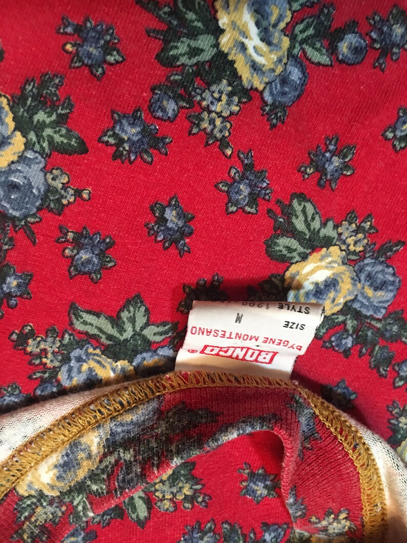 May include: A white tag with black text that reads "BONGO", "EUGENE MONTESANO", "SIZE M", and "STYLE 1908". The tag is attached to a red fabric with a floral pattern.