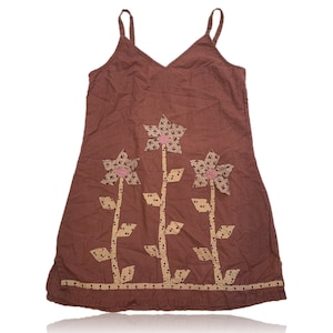 Puede incluir: Un vestido de verano marrón con tirantes finos y cuello en V. El vestido presenta tres grandes aplicaciones de flores con centros rosas y pétalos estampados. El dobladillo está decorado con un borde estampado.