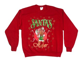 Op de afbeelding: Rode kersttrui met een groen en wit ontwerp van Santa's Helper die een zak met cadeaus vasthoudt. Het ontwerp is omringd door sneeuwvlokken en snoepstokken.