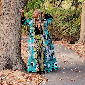 May include: A woman wearing a long, teal, black and white patterned coat with a black top and yellow and black patterned trousers. She is standing next to a large tree in a park setting.