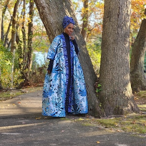 May include: A woman wearing a blue and white floral patterned coat and matching head wrap. The coat has a dark blue trim and is worn over a black turtleneck. The background features trees and a walking path.