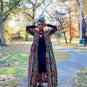 May include: A woman wearing a colorful patterned duster coat, black dress, and head wrap stands on a paved path in a park. The coat features vibrant patterns in red, blue, green, and orange. The background includes trees with fall foliage.