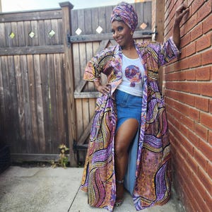 May include: A long, open-front kimono robe featuring a vibrant purple, orange, and yellow paisley-inspired pattern, worn over a white tank top with a circular graphic print and a blue denim skirt with a high front slit, paired with a matching headwrap.