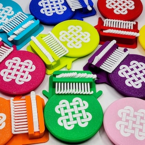 May include: A selection of brightly coloured weaving tools in shades such as blue, green, pink, orange, and purple. Each tool has a white comb-like structure and a round base with a white Celtic knot design. The tools are arranged on a white surface.