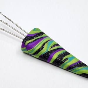 May include: A three-pronged latch hook tool with a decorative handle. The handle has a swirling pattern of black, purple, green, and blue. The metal hooks extend from the handle's top.