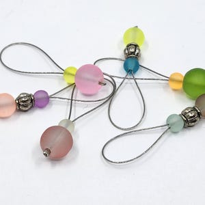 May include: A selection of colourful beaded charms. Each charm features a frosted glass bead in shades of pink, yellow, green, and blue, accented with silver-toned metal beads and wire loops. The charms are arranged against a white background.