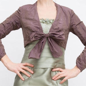 May include: A mauve cropped jacket with a bow at the front. The jacket is made of a soft, textured fabric and has long sleeves.