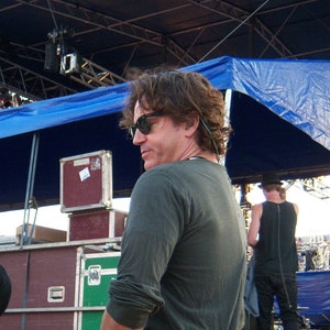 May include: A man wearing a dark gray long-sleeved shirt and blue jeans stands in front of a stage with stacked wooden crates. He is wearing sunglasses and has a microphone attached to his belt.